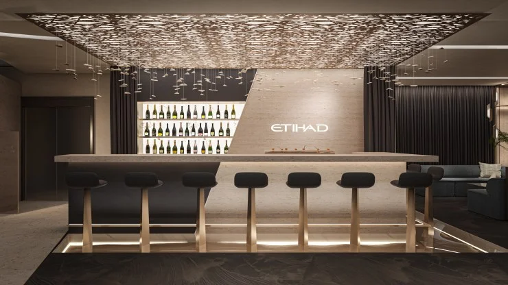 Interior of a sleek bar area featuring a modern design, with a stylish illuminated counter, high stools, and a decorative ceiling element. A wine display is visible in the background, showcasing various bottles.