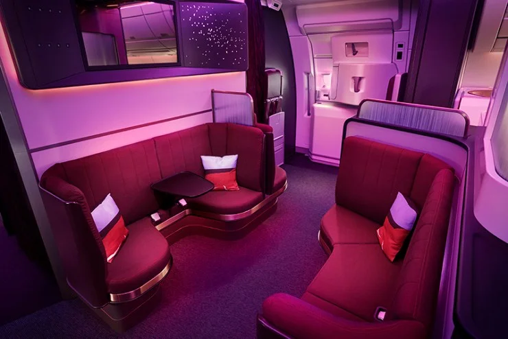 Luxurious airplane lounge area featuring two plush seating arrangements with red upholstery and decorative cushions, ambient purple lighting, and sleek design elements.