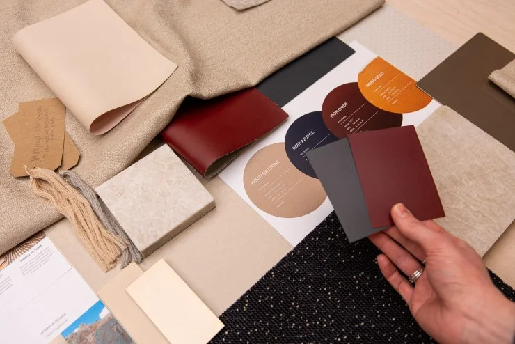 A collection of fabric swatches, color samples, and materials laid out on a table, with a person's hand holding two color cards. The arrangement includes various textures and shades, suggesting a design or decor project.