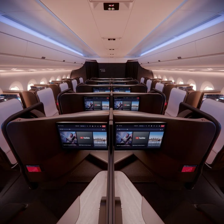 Interior view of an airplane cabin featuring spacious seating with individual entertainment screens displaying YouTube, illuminated by soft blue ambient lighting.