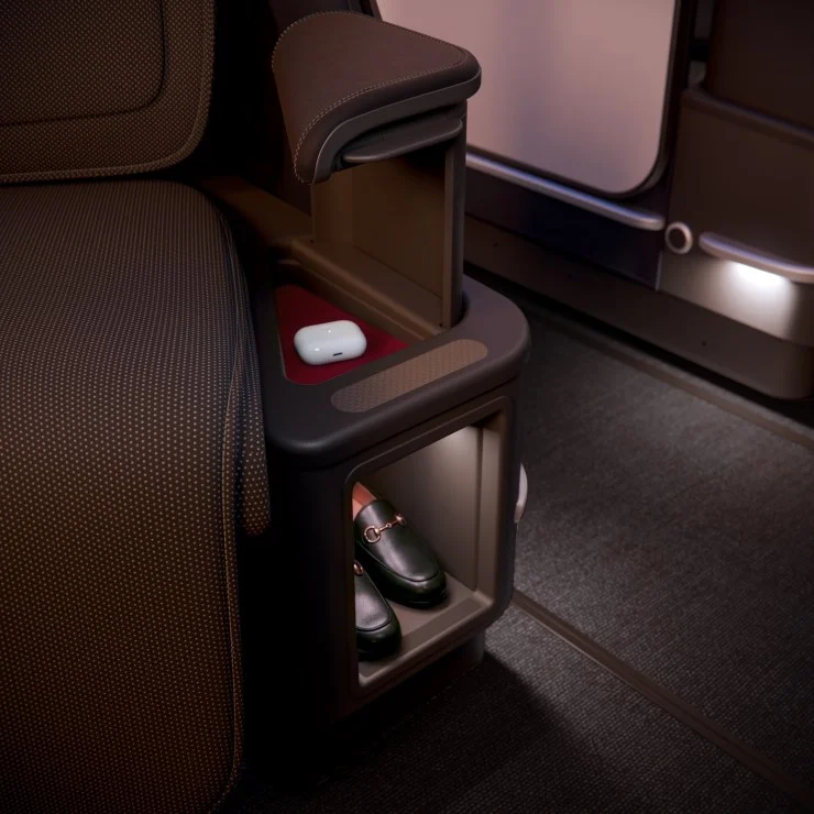 A close-up view of a luxurious airplane seat's side compartment, displaying a pair of green shoes and a set of wireless earbuds, with a textured armrest in the background.