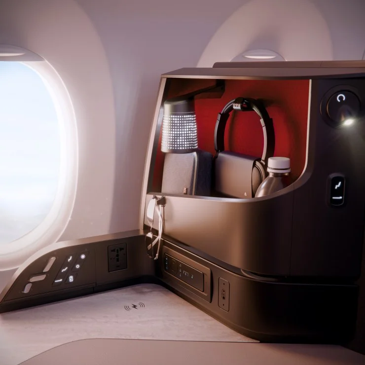 Interior view of an airplane seat with a window, showcasing a storage compartment containing headphones, a bag, a water bottle, and a lamp, along with power outlets and controls.