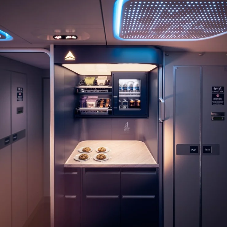 Interior view of an airplane galley showcasing a countertop with four plates of cookies, a chilled snack cabinet with assorted items, and overhead lighting.