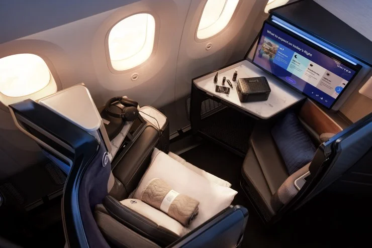 Interior view of an airplane seat with a large screen, personal amenities, and cabin windows in the background.