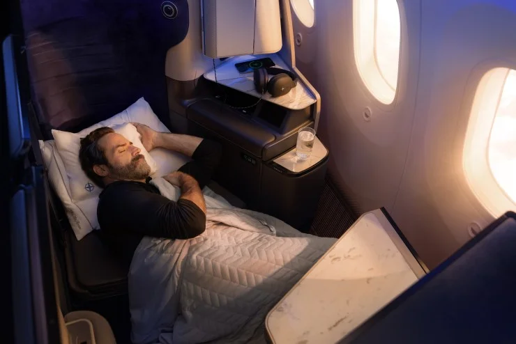 A man sleeping comfortably in a premium airplane seat, covered with a blanket, with a pillow under his head. A small table beside him holds a glass of water and headphones, while large windows show a view of the sky outside.