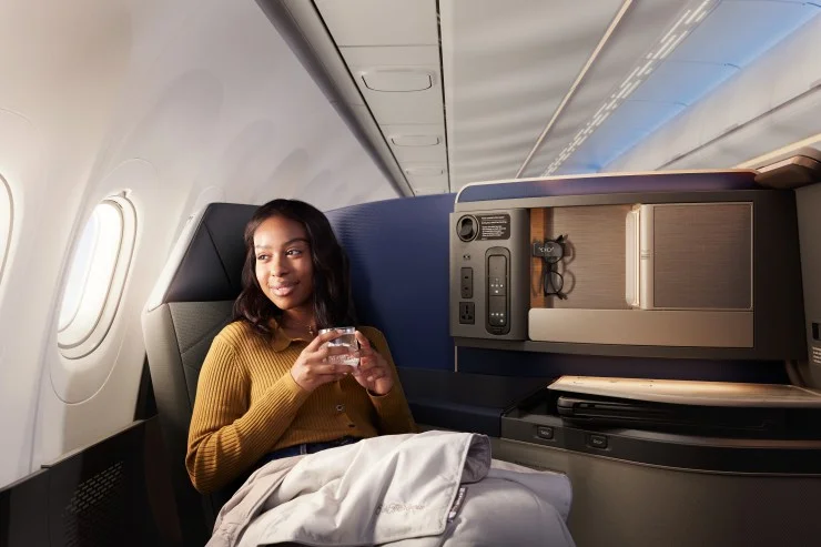 A woman sitting comfortably in an airplane seat, holding a glass of water, with modern cabin features and large windows in the background.
