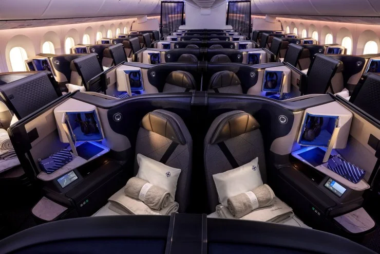 A modern airplane cabin featuring spacious seats with privacy dividers, ambient lighting, and personal entertainment systems. Each seat includes a blanket, headphones, and storage compartments.