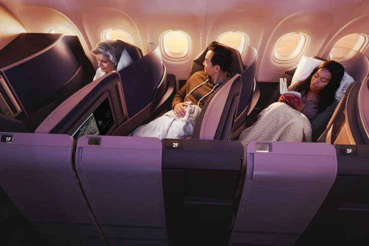Interior view of a business class cabin on an airplane, featuring three passengers seated comfortably with blankets and personal screens. One passenger is reading a book, while another is looking out the window.