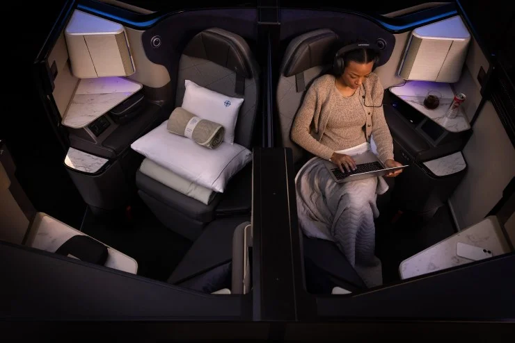 A passenger lounging in a luxurious airplane seat, using a laptop while wearing headphones, with a cozy blanket and a pillow nearby.