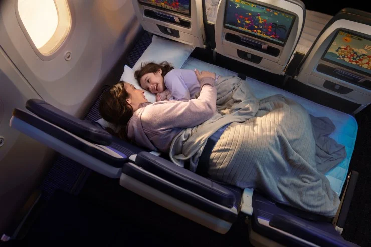A mother and daughter lying together on the floor of an airplane, covered with a blanket and smiling while enjoying each other's company.