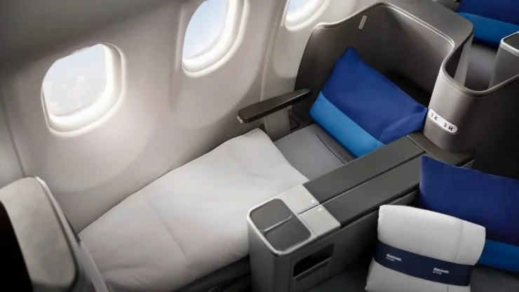Interior view of an airplane seat that converts into a flat bed, featuring blue and gray pillows and a small table, with windows revealing an aerial view.