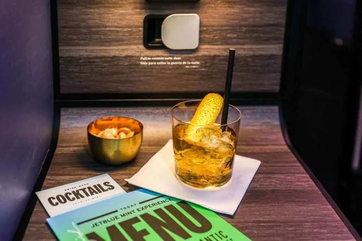 A close-up of an in-flight entertainment setup featuring a glass of cocktail with a garnish, a bowl of snacks, and a menu on a table in an airplane cabin.