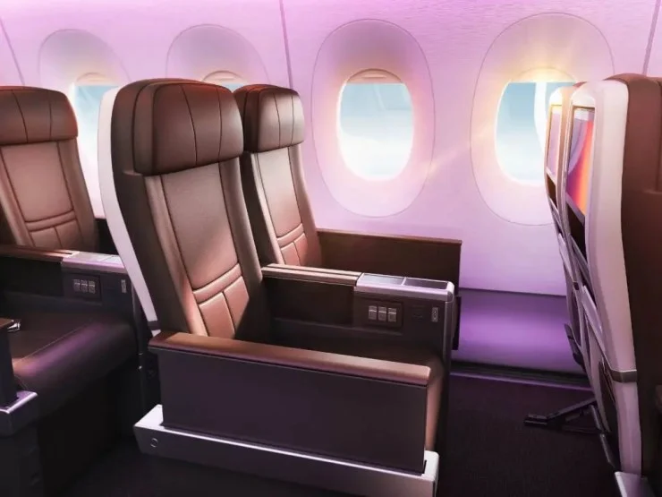 Interior view of an aircraft's premium seating, featuring plush brown seats, armrests, and side panel controls. Large windows provide a view of the outside with soft lighting enhancing the cabin ambiance.