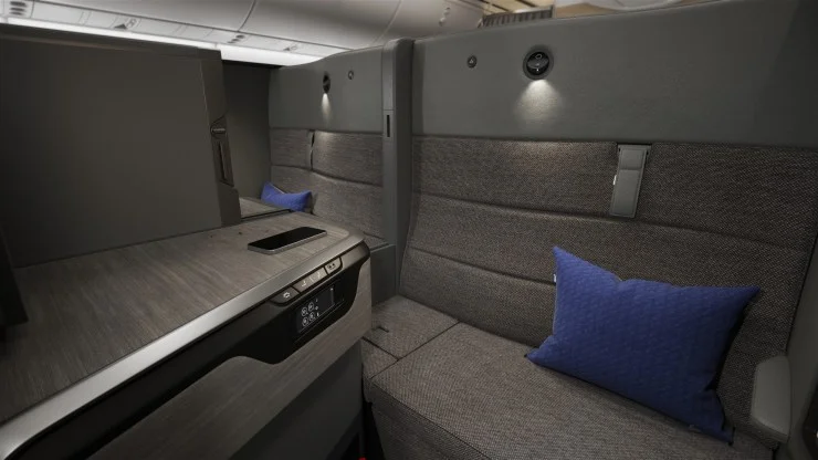 Interior view of ANA's new business class suite, THE Room FX, featuring a sleek design with gray fabric seating, a blue decorative pillow, an integrated side console with controls, and ambient lighting.