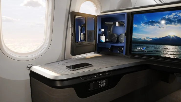 Interior view of ANA's new business class suite, featuring a spacious console with a phone placed on it, an entertainment screen displaying a scenic view, and storage compartments holding headphones and amenities.
