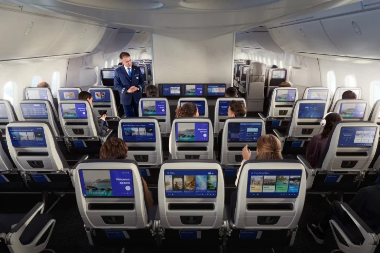 Interior view of a United Airlines aircraft featuring multiple rows of economy seats, each equipped with personal entertainment screens. A flight attendant stands at the front, greeting passengers.