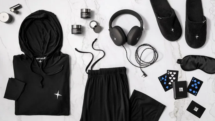 A flat lay arrangement showcasing United Airlines' premium amenities including black hoodie, pants, noise-canceling headphones, slippers, skincare products, an eye mask, and playing cards.