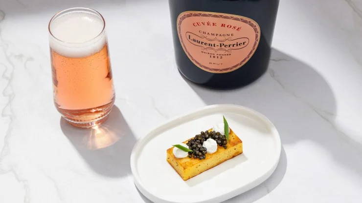 A glass of Laurent-Perrier Cuvée Rosé Champagne accompanied by a gourmet amuse-bouche featuring caviar on a rectangular appetizer with herbs, set against a marble background.
