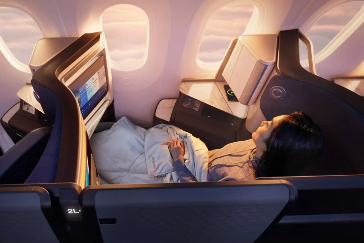 A passenger relaxing in a United Airlines Polaris Studio business class seat on a Boeing 787-9 Dreamliner, with a blanket and a personal entertainment screen, while looking out of the window.
