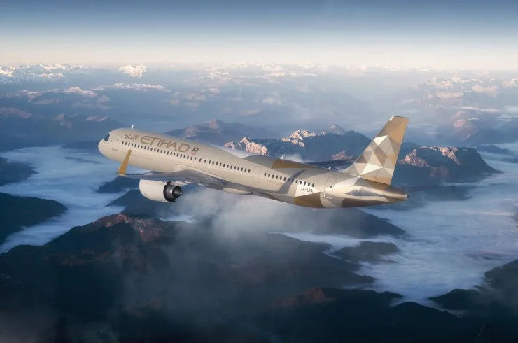 An Etihad A321LR aircraft flying over a mountainous landscape, showcasing the airline's distinctive livery against a clear blue sky.